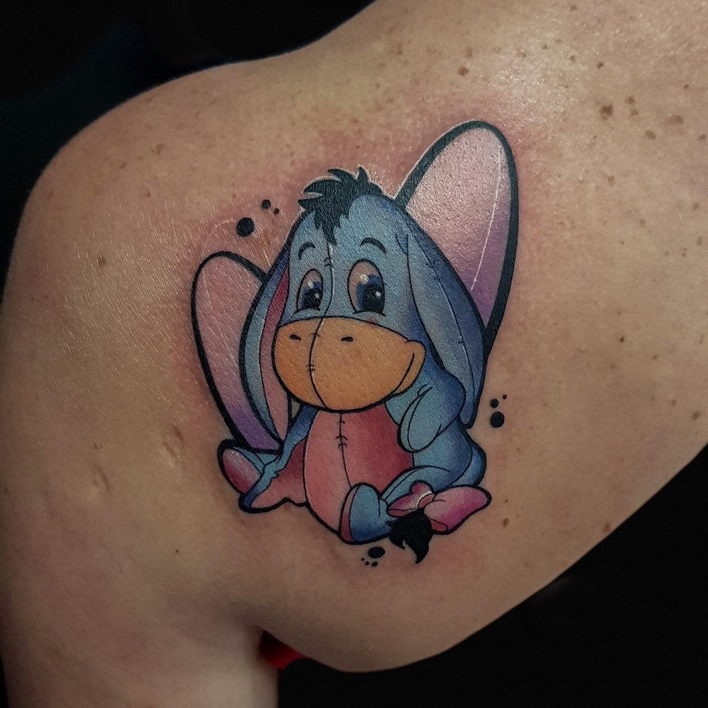 Colorful tattoo of a cute, sitting Eeyore from Winnie the Pooh on a person's shoulder